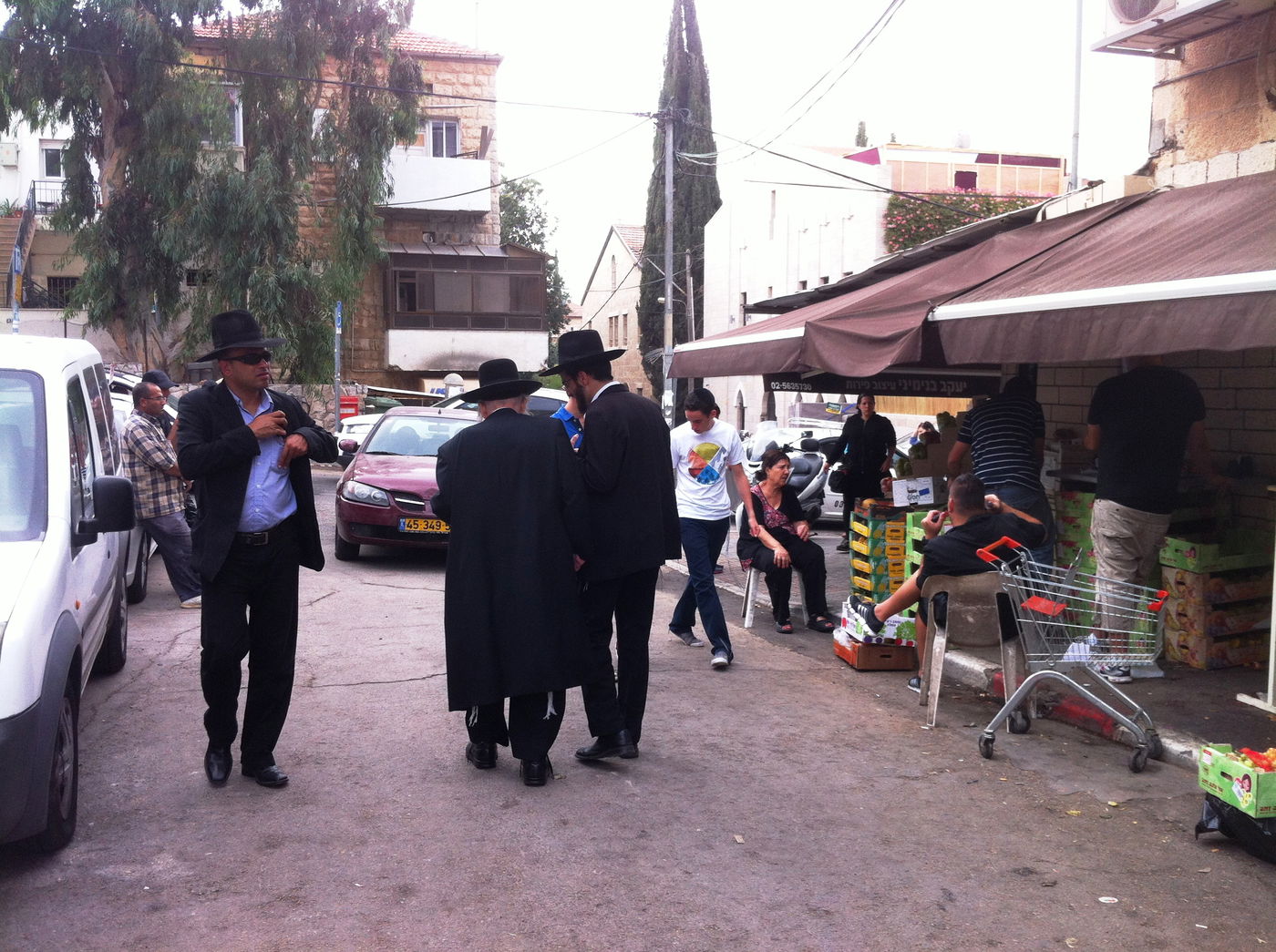 ,,והנה רגע כזה של שעת צהריים ב"שערי חסד": מימין, ליד חנות הפירות, צועד נער בטישרט וציציות בחוץ. משמאל הולך אמריקאי במשקפי שמש, חולצת תכלת וחליפה. ובמרכז - ר' שמואל אוירעבך פוסע לאט לאט, מהישיבה אל ביתו הפרטי, שקוע בשיחה עם תלמיד. צילום סלולרי: ידידיה מאיר