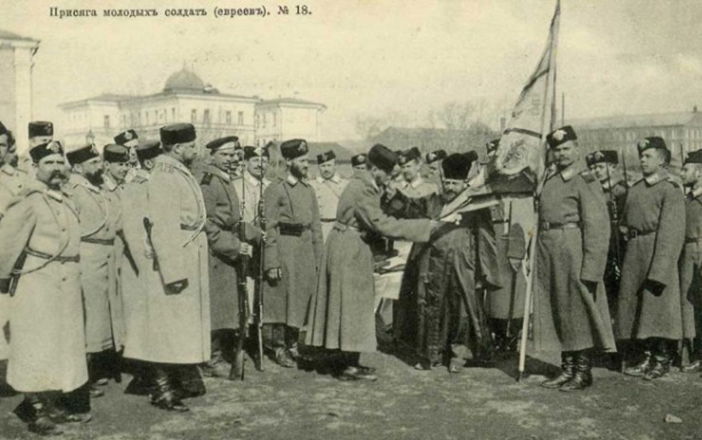בספר תורה ובדגל - טקס השבעת טירונים יהודים לצבא האימפריה הרוסית (צבא בית רומנוב), ראשית המאה ה-20. במרכז התמונה נראה חייל נשבע אמונים על ספר תורה או תנ"ך בפני רב, בנוכחות נושא דגל היחידה